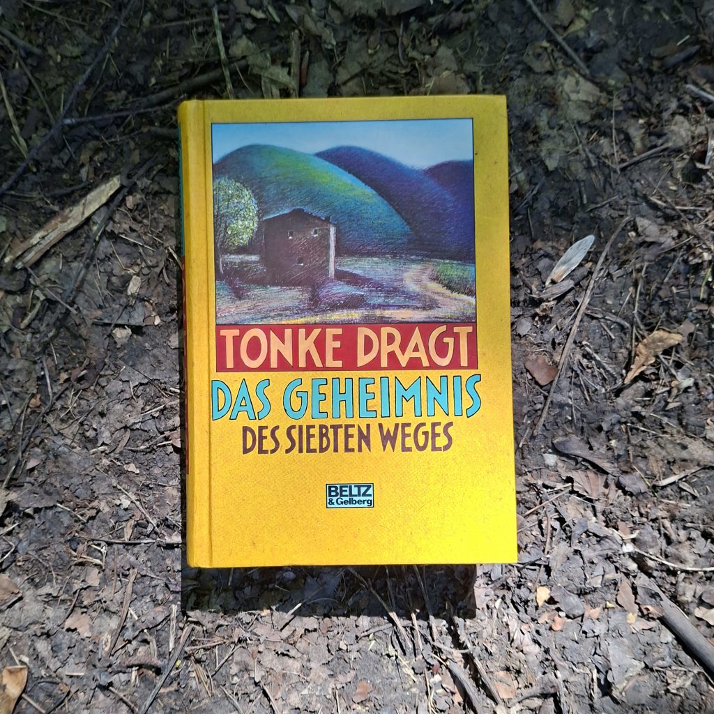 Buchcover von Das Geheimnis des siebten Weges von Tonke Dragt, erschienen im Verlag Beltz & Gelberg. Das gelbe Cover zeigt ein kleines Haus vor grünen Hügeln, fotografiert auf Waldboden mit Blättern und Zweigen.