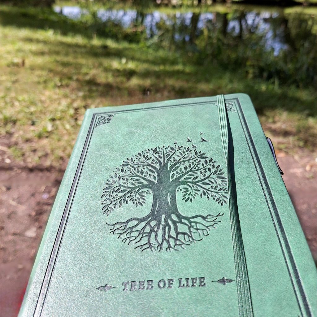 Grünes Notizbuch mit einem Baum-des-Lebens-Motiv am Ufer eines Sees als Symbol für Geschichten, Kreativität und Lesefreude.