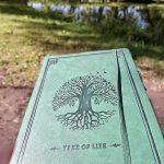 Grünes Notizbuch mit einem Baum-des-Lebens-Motiv am Ufer eines Sees als Symbol für Geschichten, Kreativität und Lesefreude.