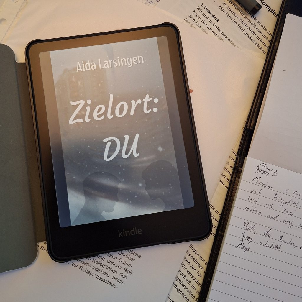 Foto: Ein Kindle E-Reader mit dem Cover des Romans „Zielort: DU“ von Aida Larsingen (dunkle Silhouetten am Fenster) liegt auf einem Stapel Papiere, daneben handschriftliche Notizen.