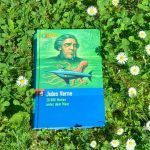 Buch „20.000 Meilen unter dem Meer“ von Jules Verne liegt auf einer grünen Wiese mit vielen weißen Gänseblümchen im Sonnenlicht.