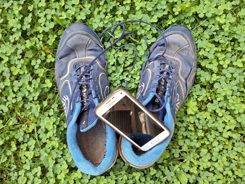 Ein Paar abgenutzte blaue Sportschuhe liegt auf dichtem, grünem Klee. Zwischen den Schuhen befindet sich ein weißes Samsung-Smartphone mit schwarzem Bildschirm.