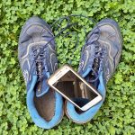 Ein Paar abgenutzte blaue Sportschuhe liegt auf dichtem, grünem Klee. Zwischen den Schuhen befindet sich ein weißes Samsung-Smartphone mit schwarzem Bildschirm.